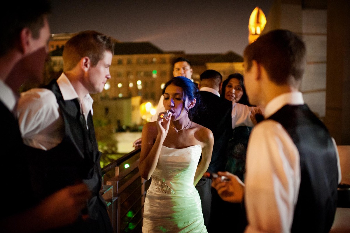 beautiful bride smokes a cigar at a reception as groom and friends watch