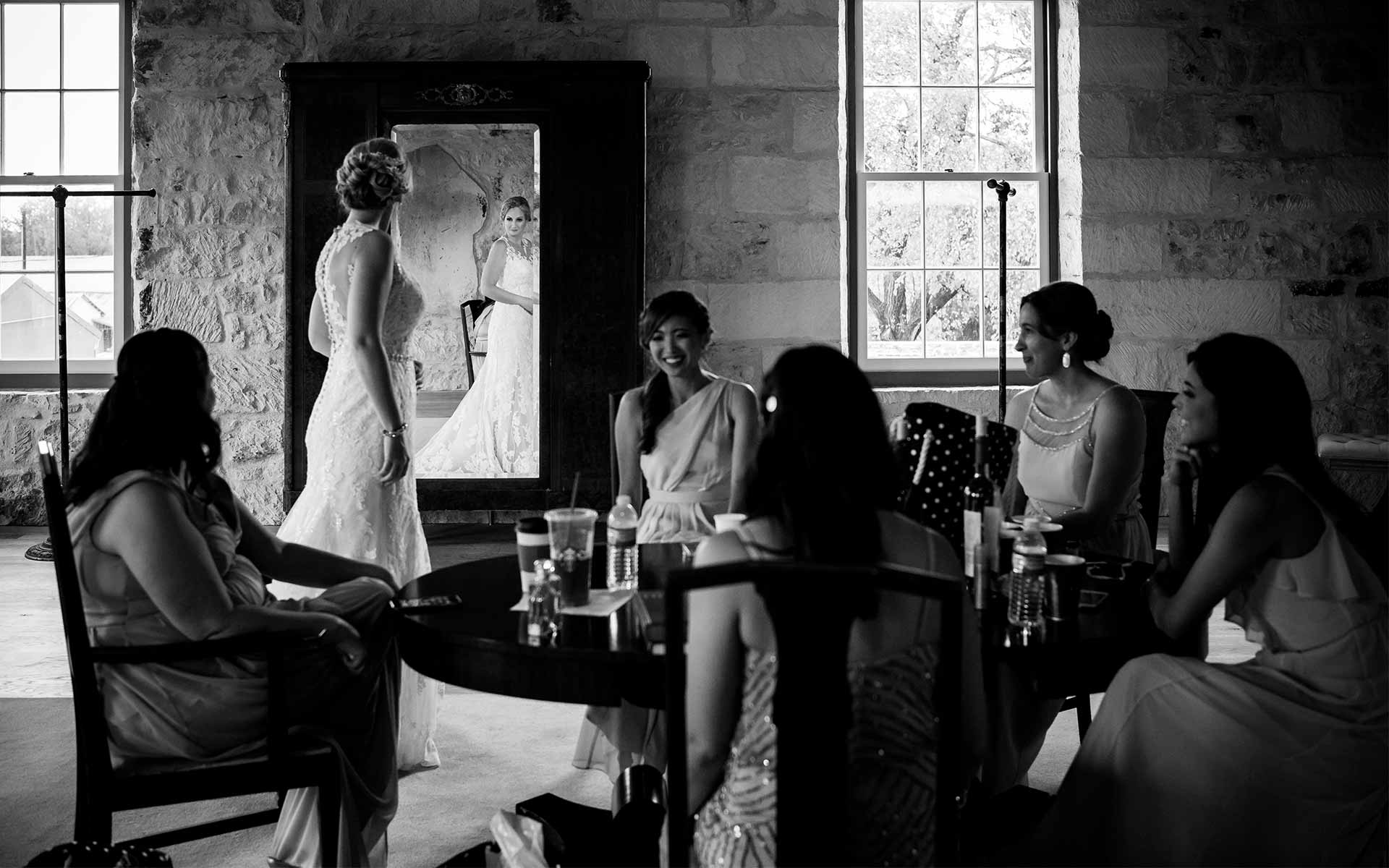 Bride steals a glance in the mirror as bridesmaids sit around a table at Ingenhuett on High, Comfort, Texas.
