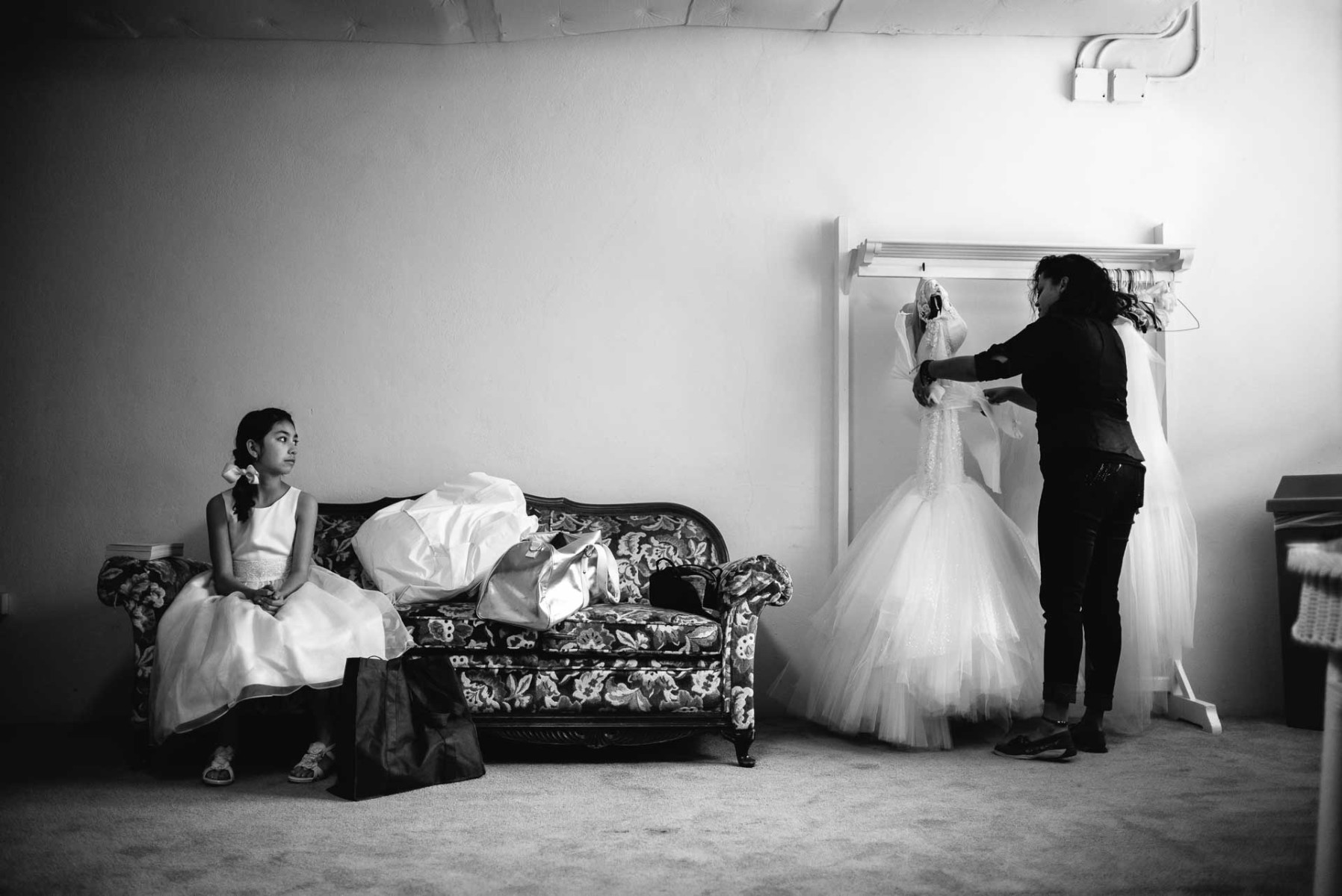 Flower girl watches her mother as the brides dress hangs on a rail pre wedding ceremony