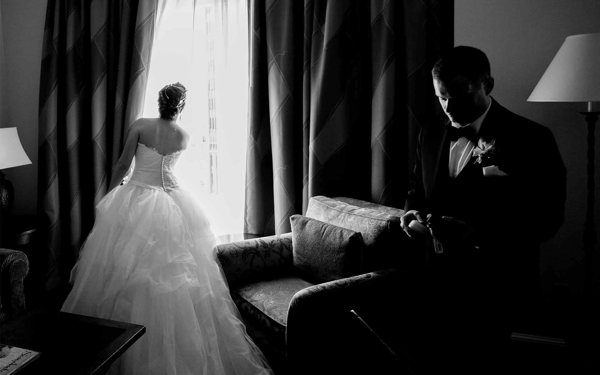 Bride steals a glance out of window pulling on curtain and groom check the time. An international award winning image.San Antonio Wedding Photographers