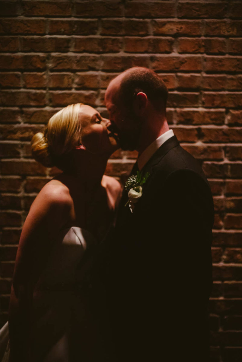 Couple kiss under a over head spot light against a brick wall at a wedding reception Brennan's of Houston, Texas.
