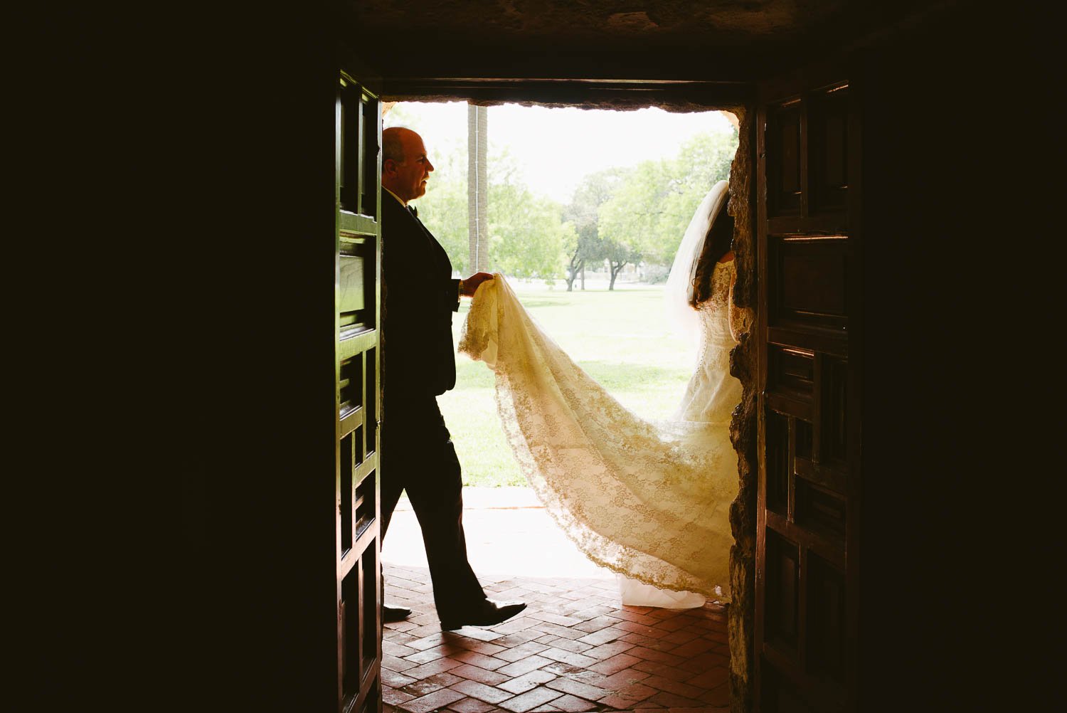 Father of the bride holds her train as they make their way to the front entrance and the beginning of the processional at Mission Concepcion Wedding-Leica photographer-Philip Thomas Photography