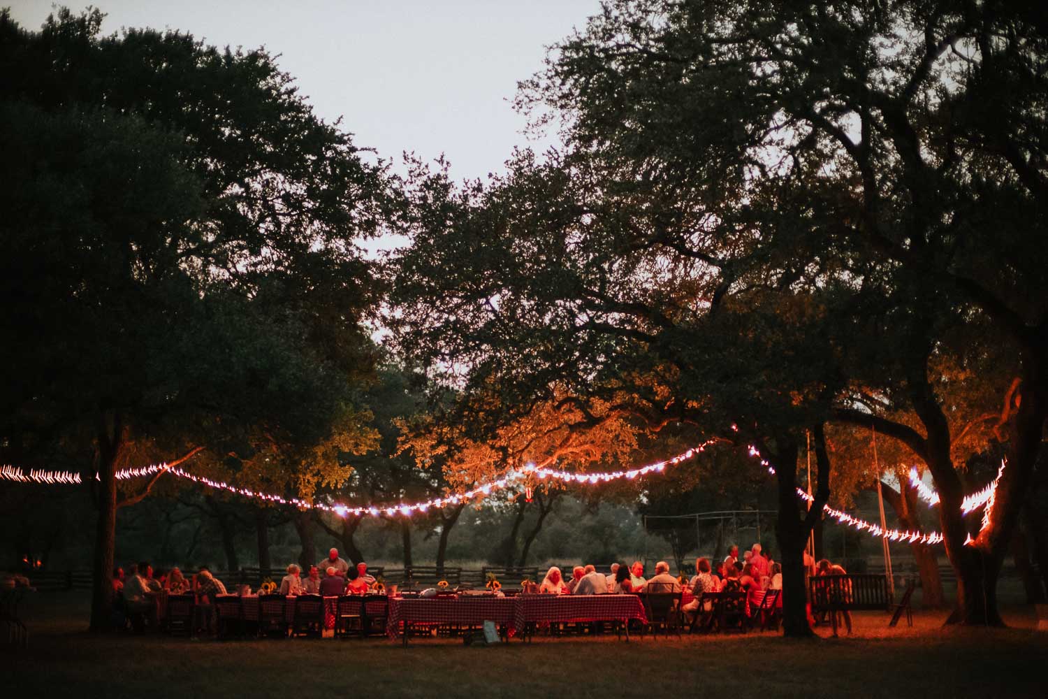 Twilight image of an outdoor rehearsal dinner party with twinkling lights and long outdoor tables