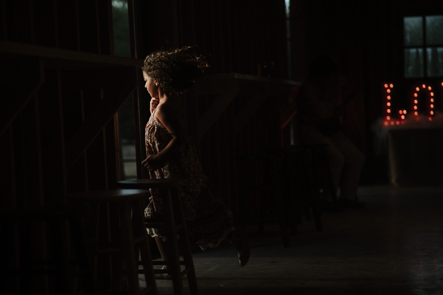 A little girl runs though a shaft of light at a barn wedding reception