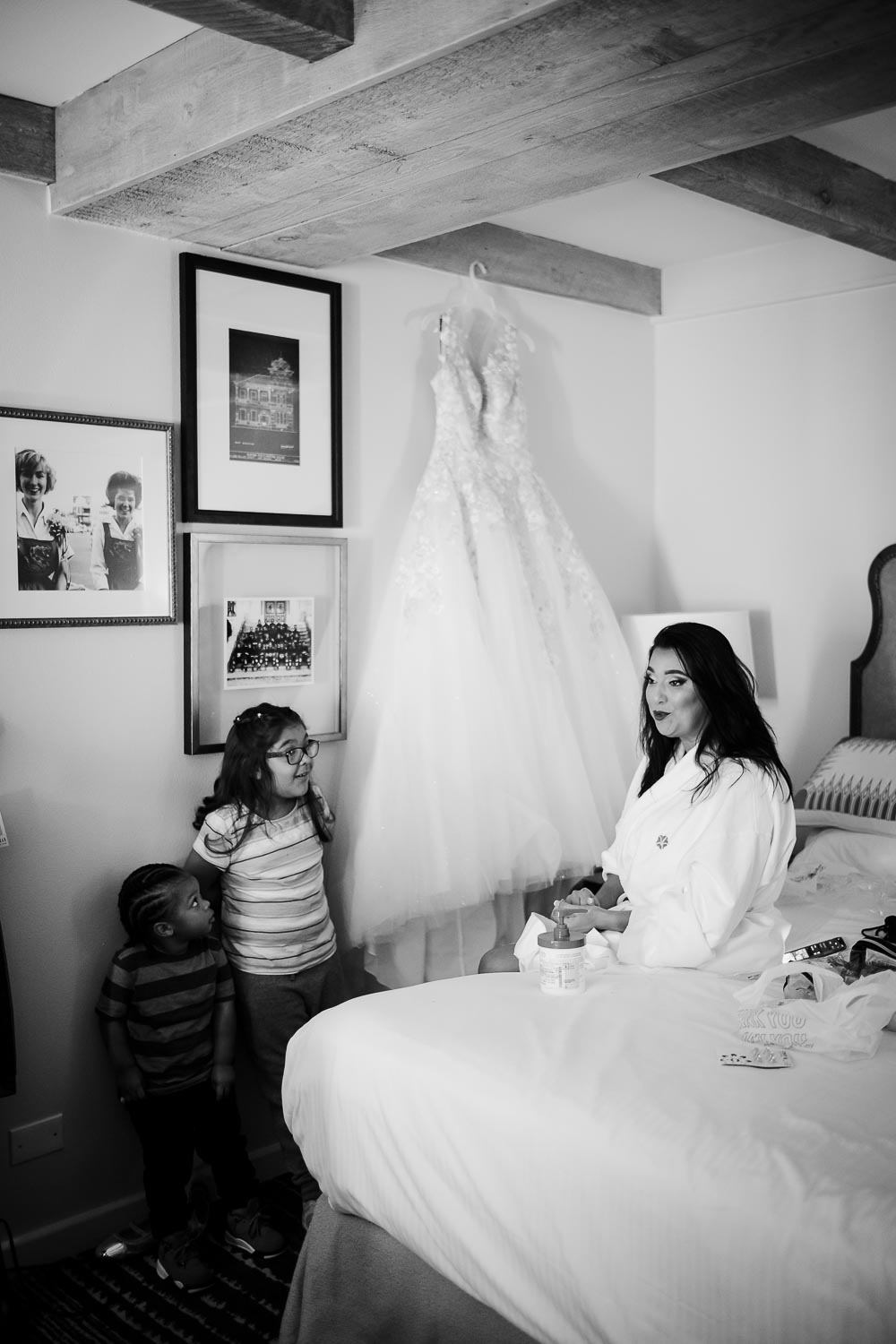 Bride sits on bed with wedding dress hanging bejind her as ring beaer and flower girl look at bride