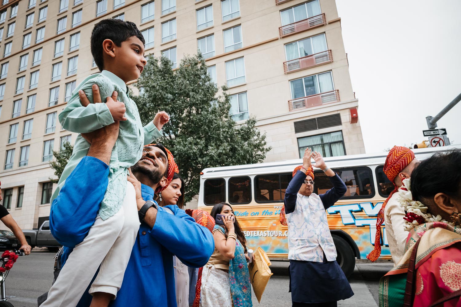 A boy sits on his father shoulders during the Baraat Brazos Hall Austin South Hindu asian wedding ceremony