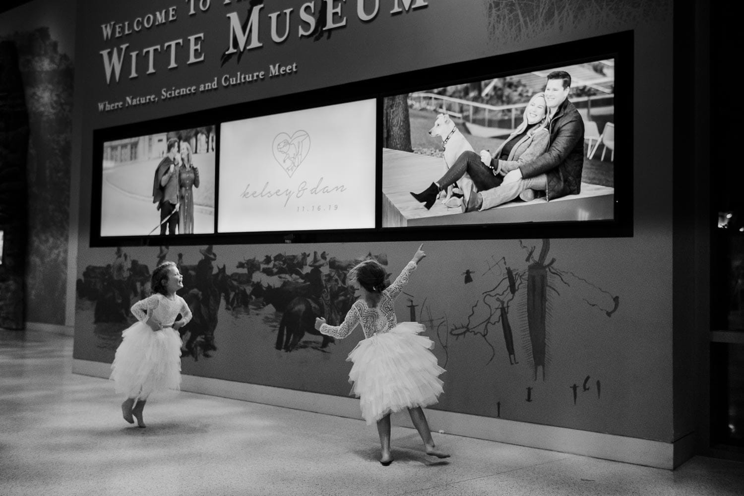 Flower Girls dance and point at a picture of the newly married couple on a large tv monitor at The Witte Museum