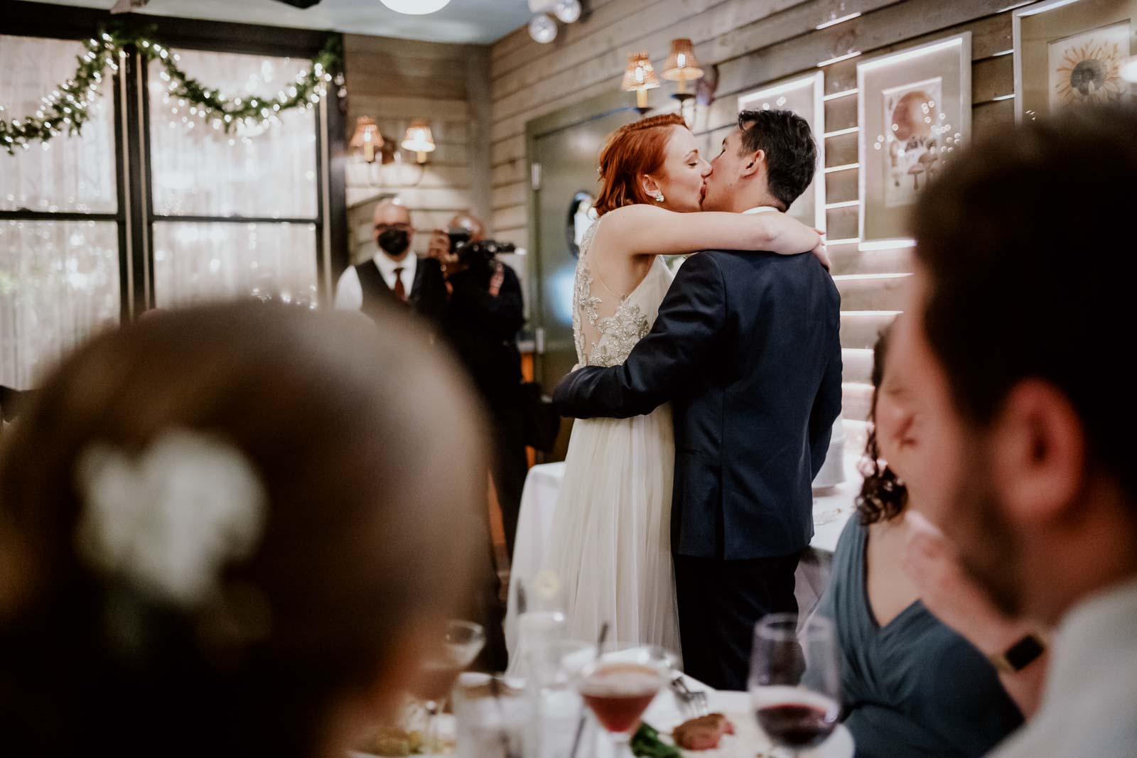 The couple kiss and embrace at Ouisie's Table wedding reception in Houston captured between guests at their tables