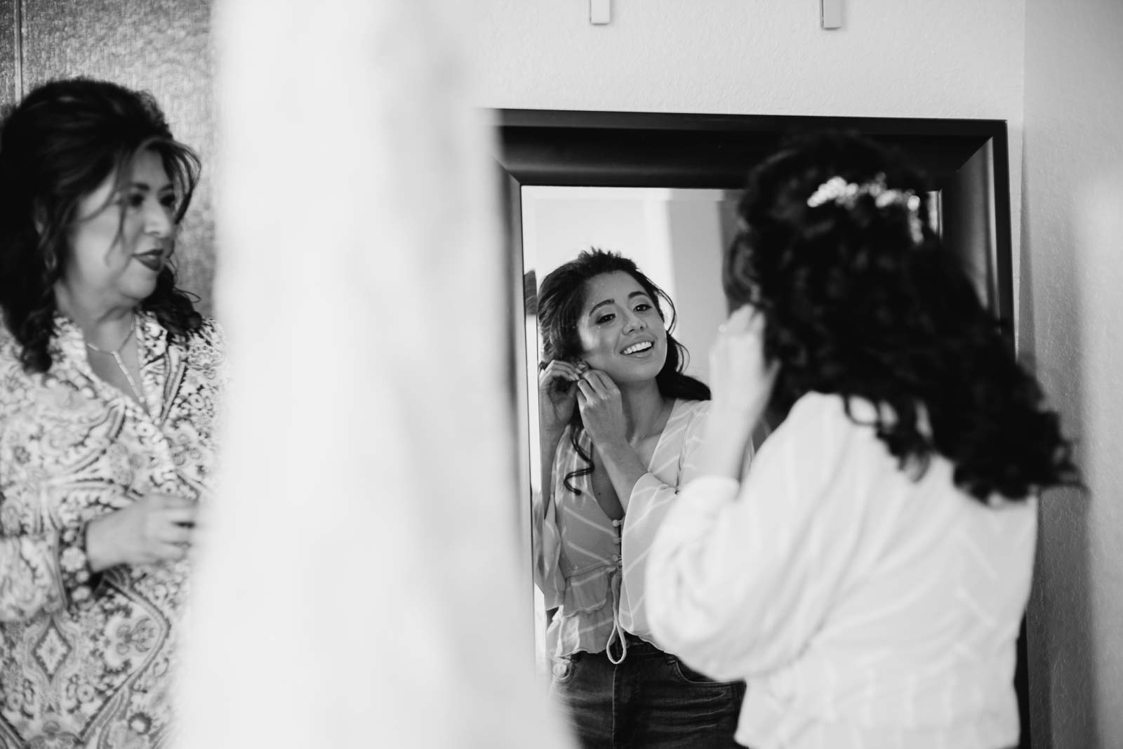 The bride looks in the mirror as she adds her earrings with the dress hanging from the ceiling and her mother looking on adoringly