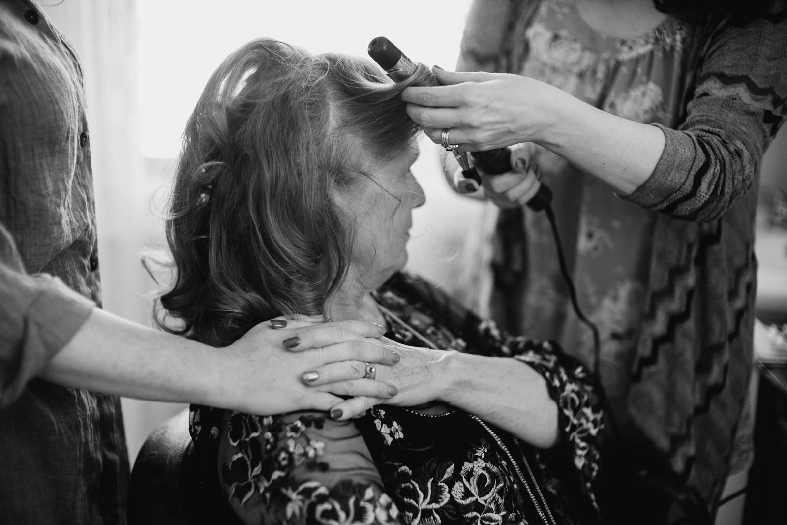 The mother of the bride and her daughters hold hands as she has her hair styled