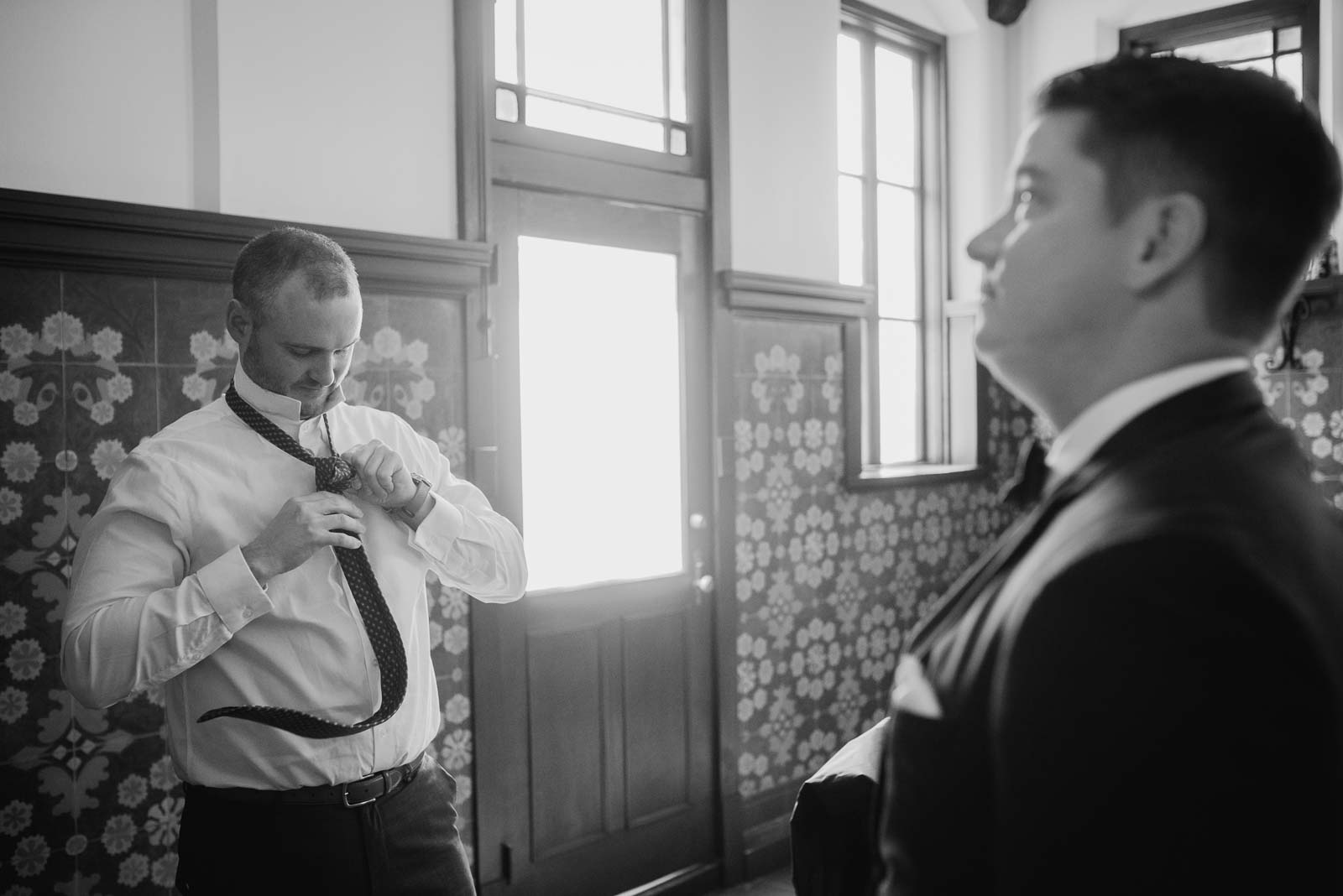 A groomsman put his tie on as the groom looks on