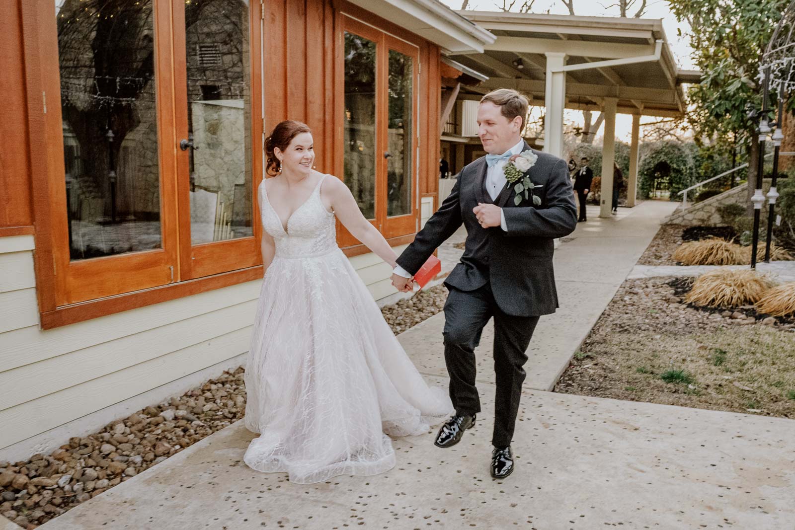 A couple trot and jump in the air in a funny moment as they leave or exit the wedding venue with family members in the background in the late winter wedding