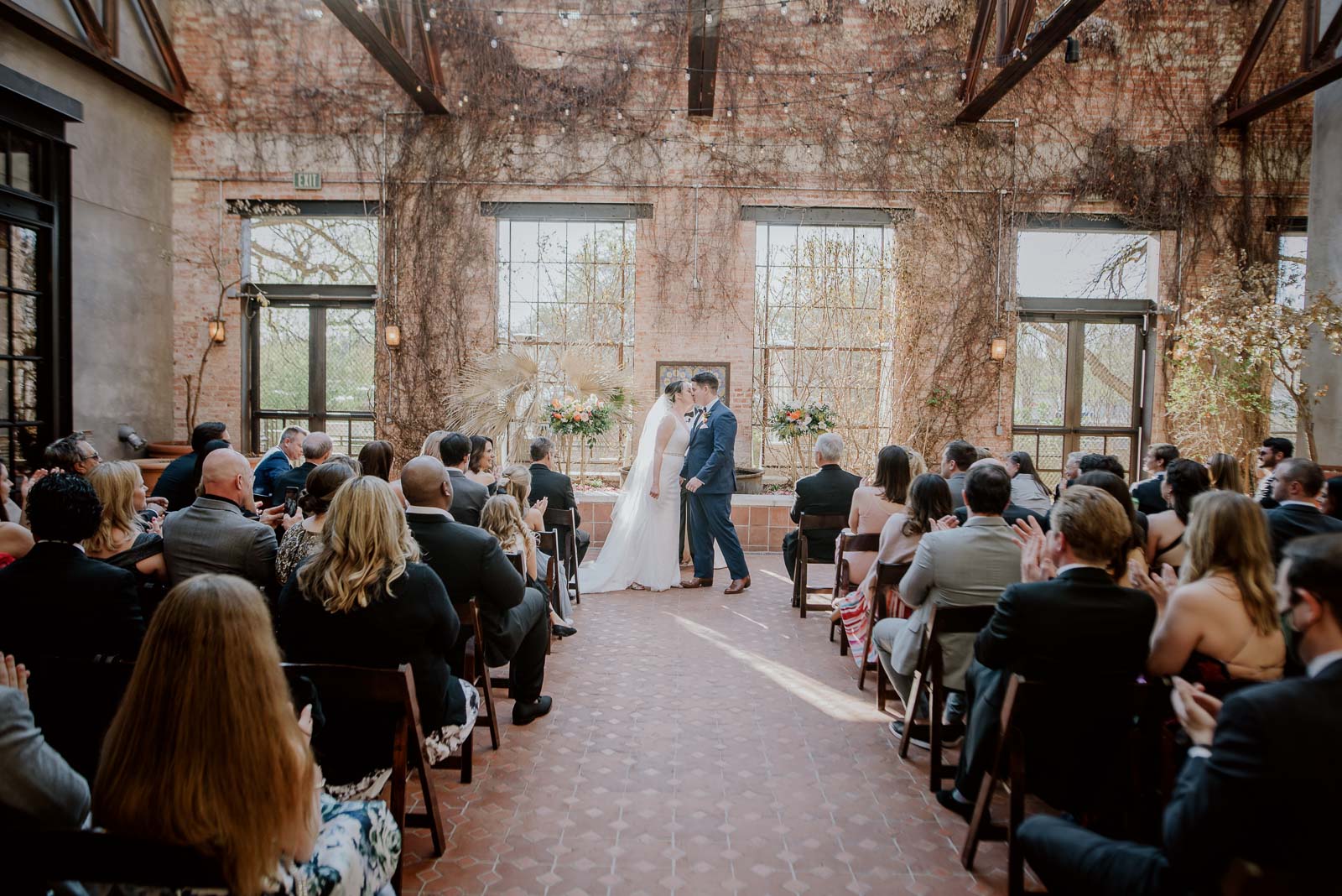 A wide angle shot photograph on the back of the wedding ceremony at Hotel Emma as a couple kiss and friends and family applaud