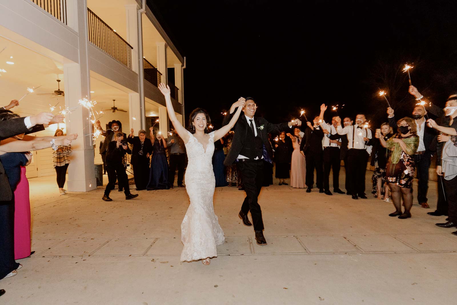 The bride and groom raise their hands in jubilation as sparklers are out up in the air by guests on their exit at the wedding venue