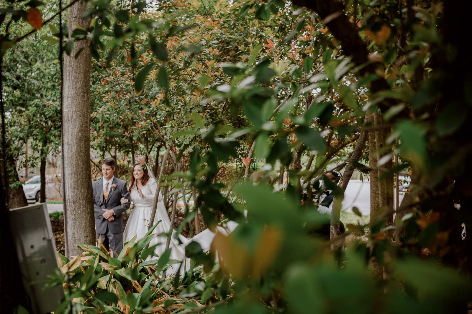 With greenery in foreground the bride is captured waving to onlookers with her father just said she’s about to walk down the aisle