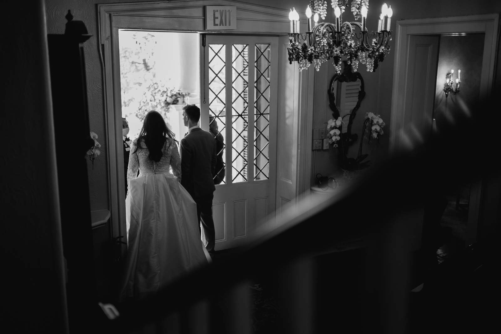 The bride and groom get ready to exit out onto the reception with the crowd waiting photographed in black-and-white for the staircase photograph with a Summilux 35mm on a lake and rangefinder through the glass pane door the bottom room hold hands