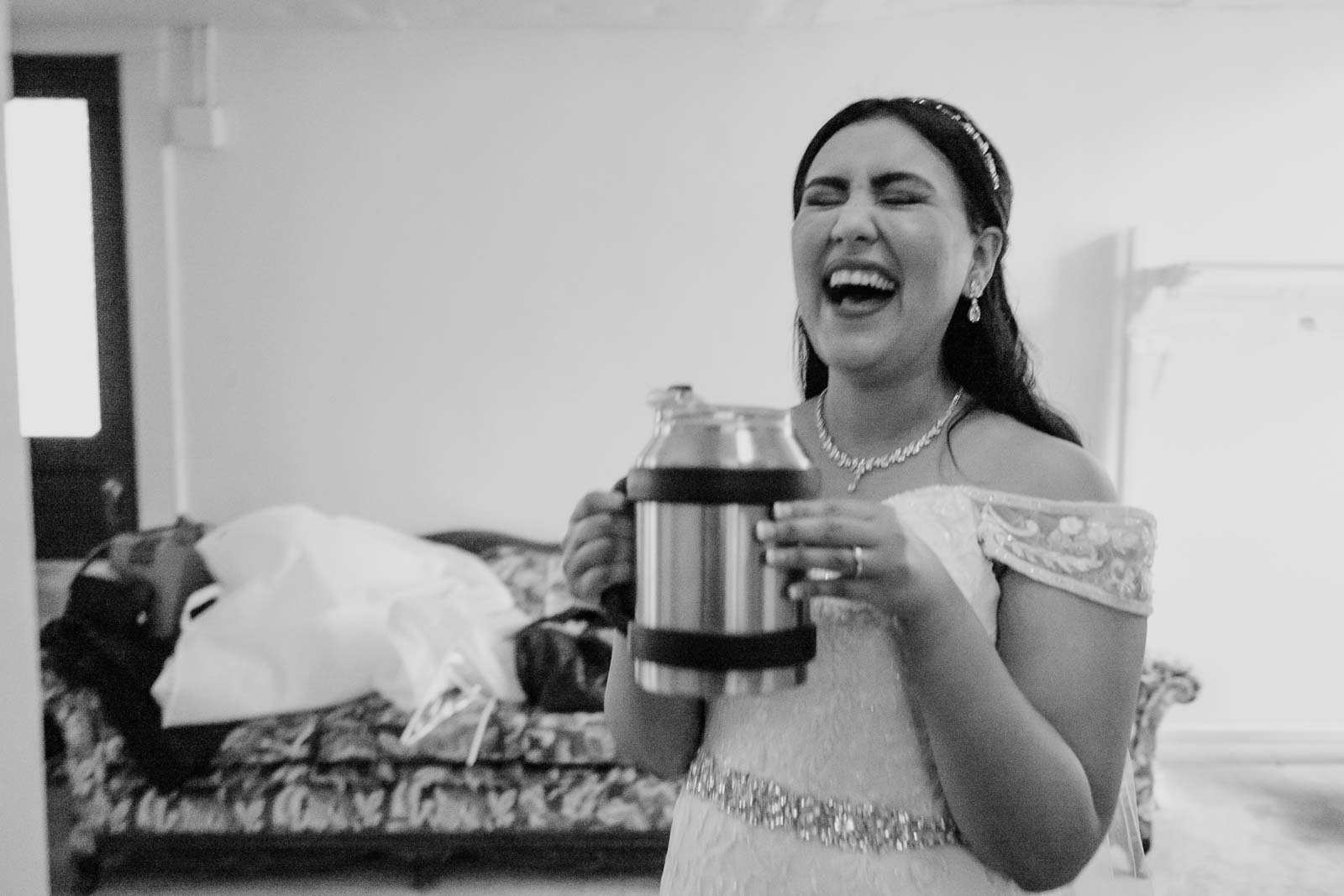 A moment of laughter has bride sips a drink from her drink carrier A best friend hold the wedding veil high so as not to wrinkle it The mother clutches the end of the veil The bride beams with a wide smile at mirror A dragged shutter images black and white leica has friends embrace at wedding Arsy the bride checks her earrings look perfect in mirror Jewellery is added to brides wrist by her mother The bride slips on her wedding shoes at With a hekping hand from her mother the bride gazes out of window The groom arrives with palm tree in background photographed through window A beautiful dress hangs from the ceiling at Details of the veil in black and white Sacred Heart Chapel-Leica photographer-Philip Thomas Photography