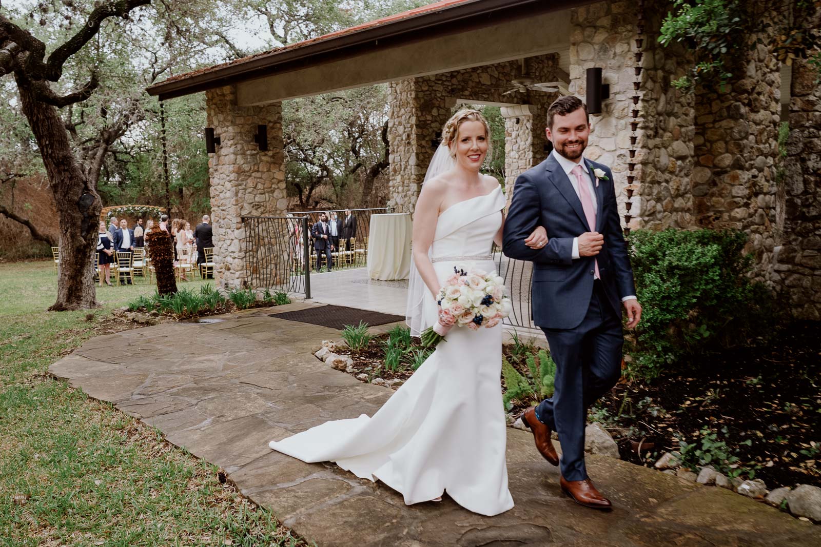 Just married couple walk away from the ceremony site at the veranda with big happy smiling faces as a bride holds up bouquet clutching the arm of the groom