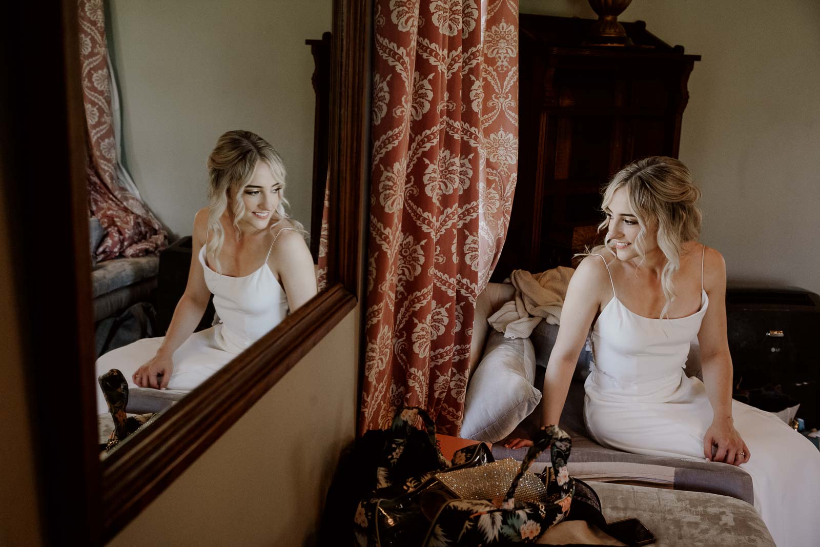 The bride sits at a window looking out and a reflection of a mirror