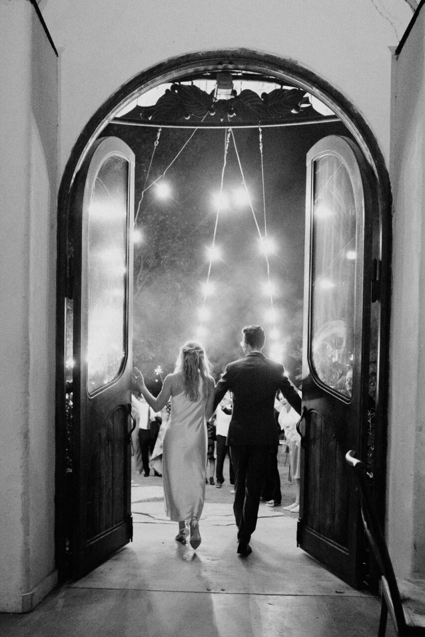 Anna and Caleb open the ballroom doors photographed from behind in glorious black and white with string lights above at Barr Mansion