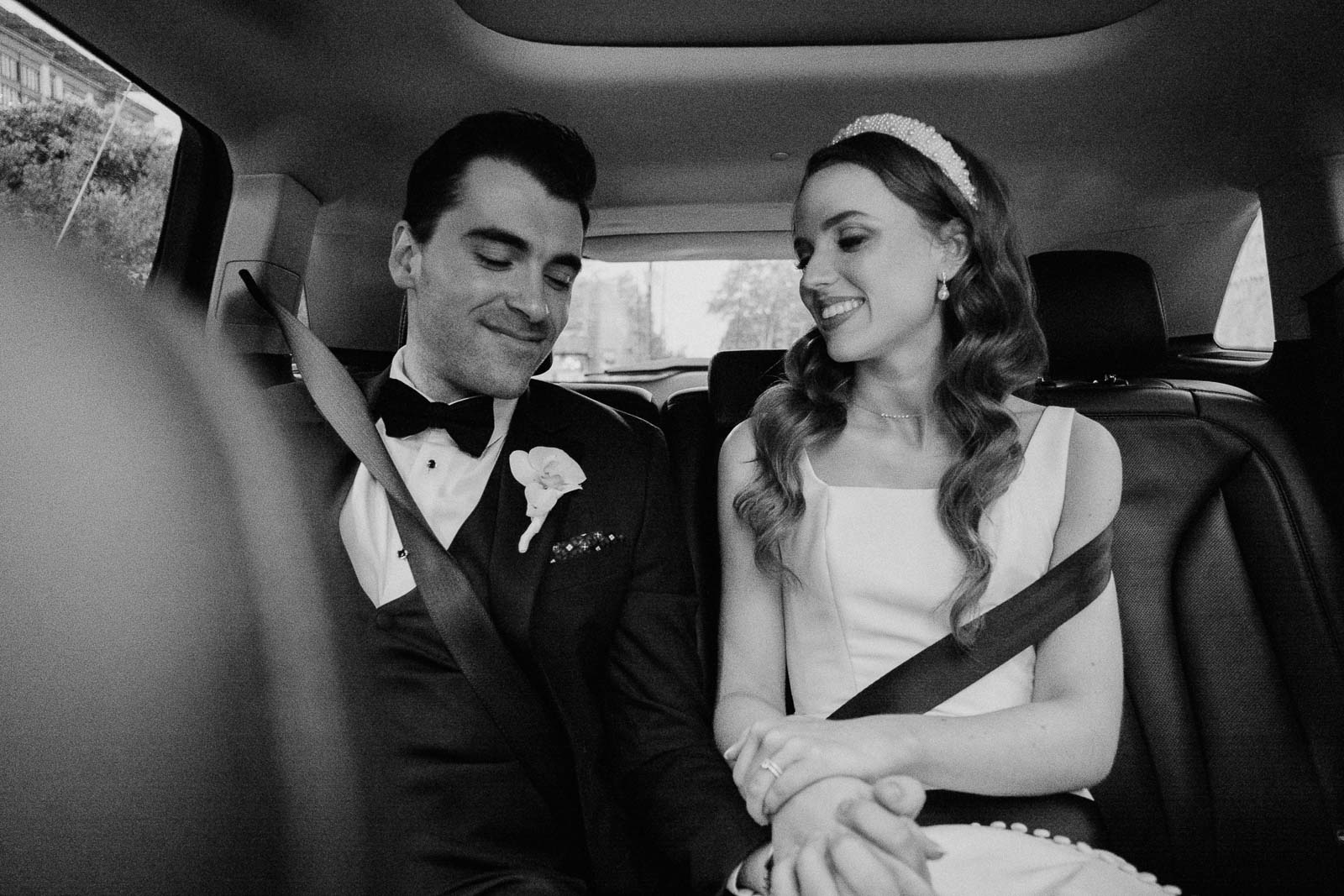 In the back of a hired SUV the newly married couple hold hands as the groom looks down to the ring