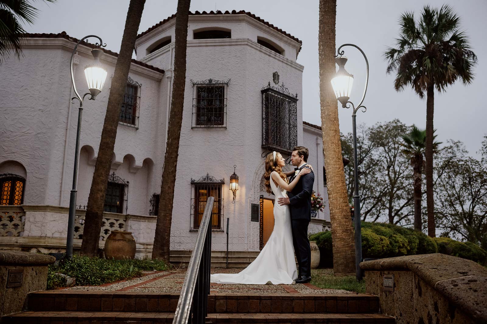 Under the lamp light a couple stand at dusk with the McNay Art Museum behind them