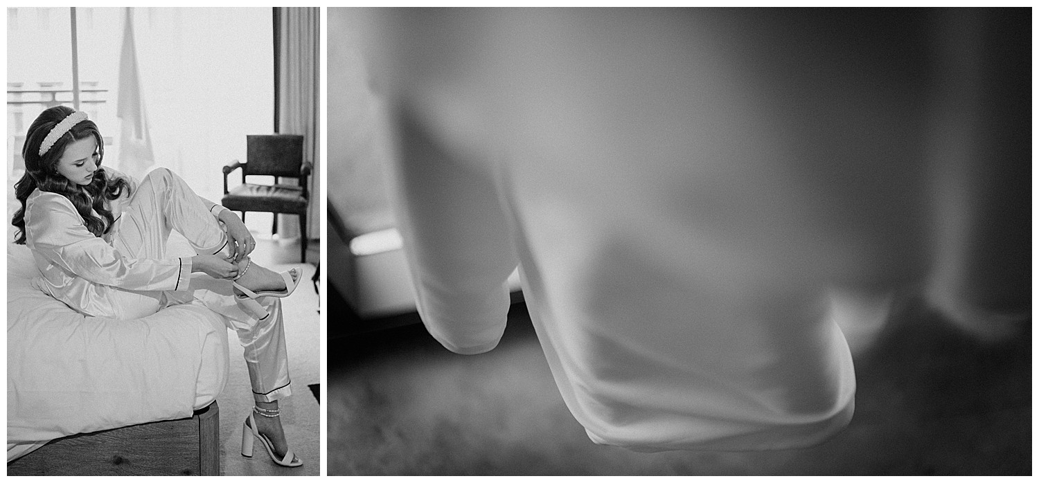 The bride slips on her shoe in her hotel room with dress hanging in the background