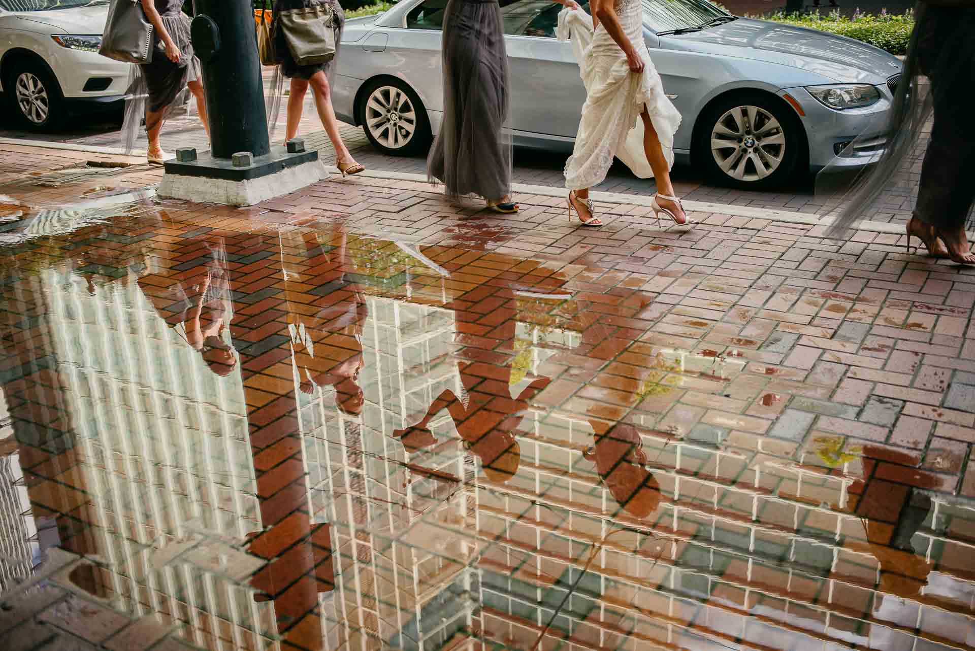 A captivating candid moment of a wedding party walking past a puddle, beautifully reflecting bridesmaids and the bride in high heels. Captured with a Leica M (Typ 240) and Summilux-M 35mm lens, this street-style wedding photo showcases elegance, symmetry, and urban charm.