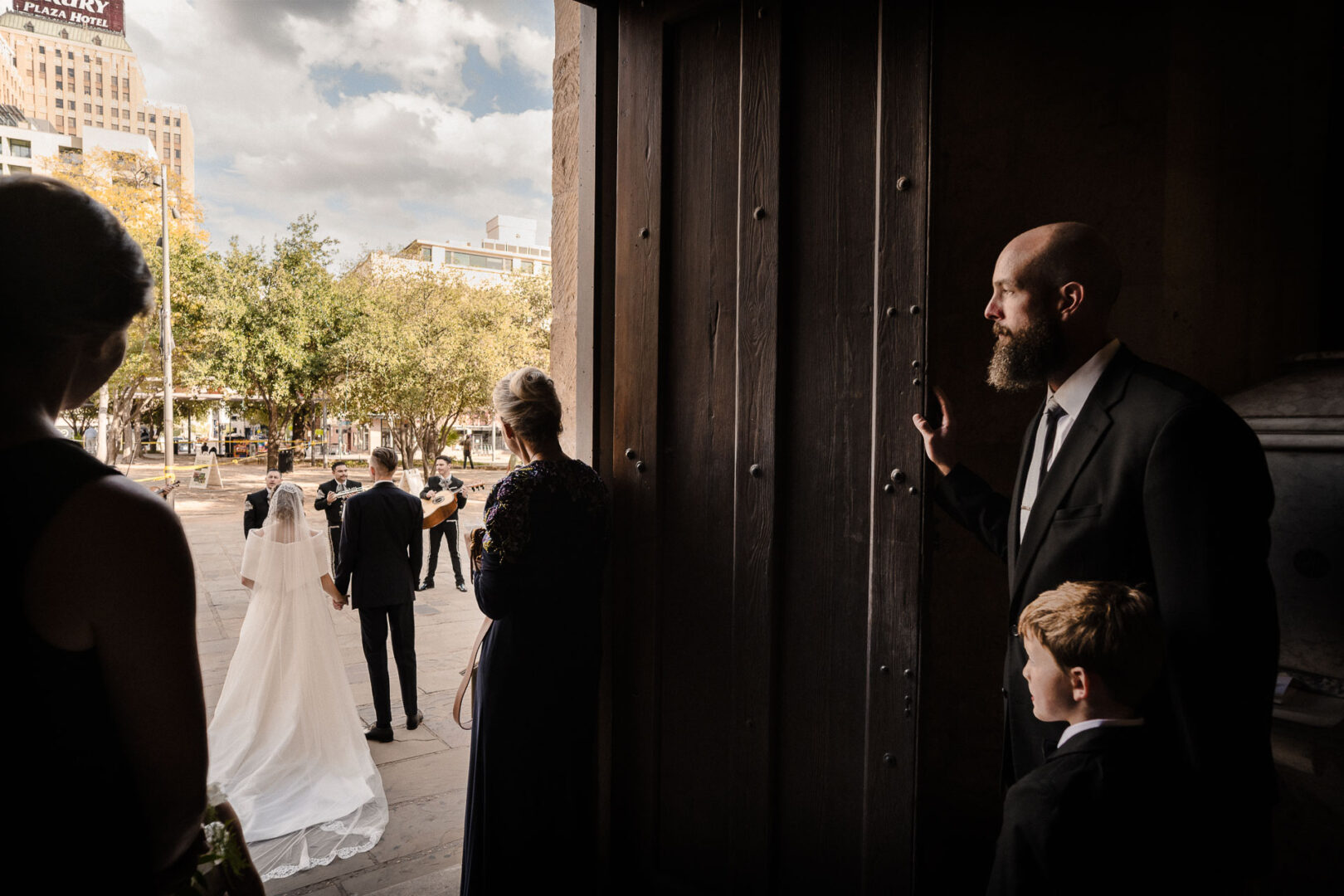 Picture of the Month: San Fernando Cathedral Wedding Moment in San Antonio 2 Bride and groom walk out of a dark church doorway into bright daylight as mariachi musicians wait outside, while a man and young boy watch from the shadows.