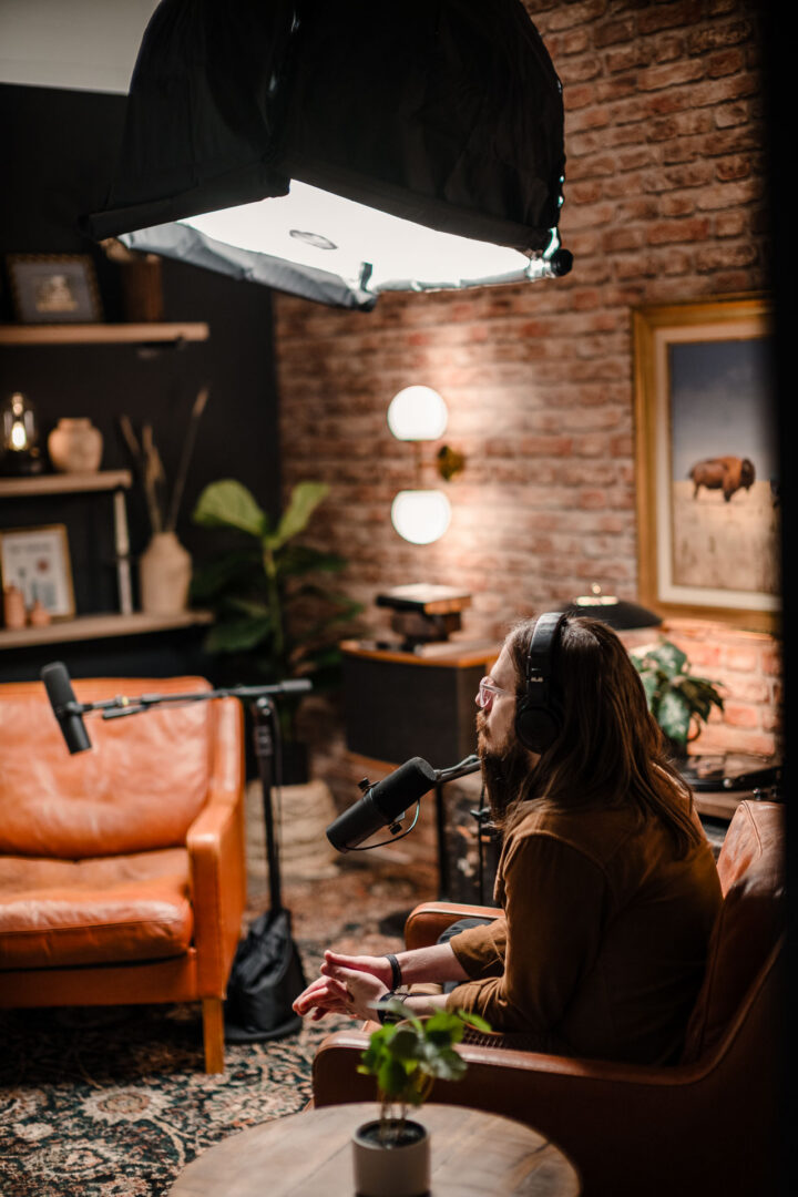 Behind-the-scenes production still showing interviewer in the foreground, talent on set, and overhead key light in a controlled studio interview setup.