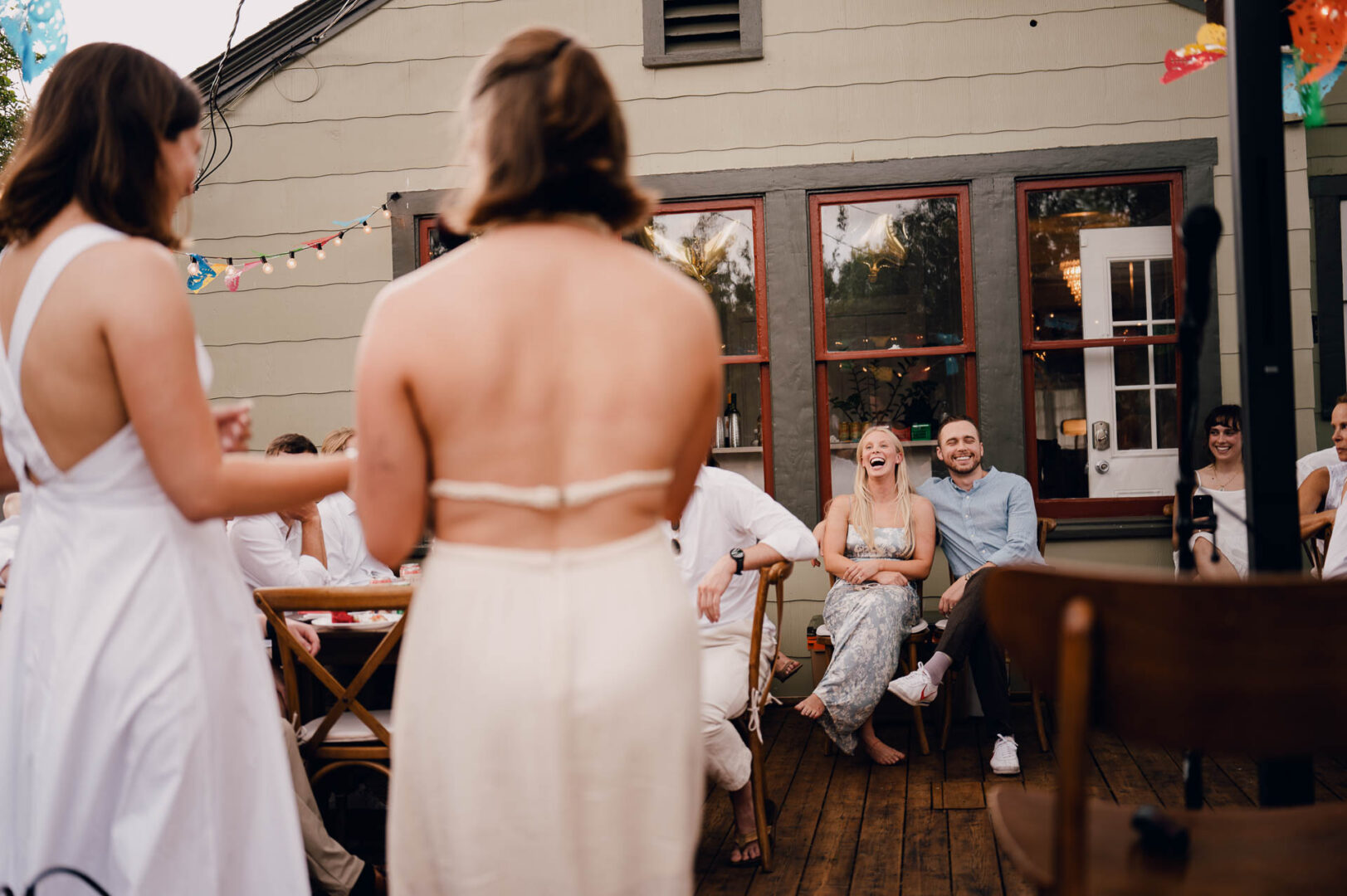 Wedding guests seated outdoors, smiling and reacting during speeches at a backyard reception.