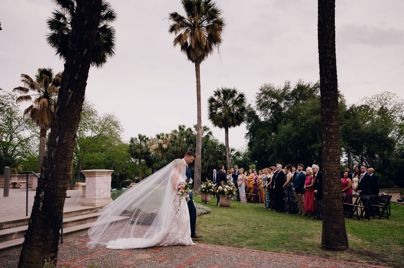 Bride walking down the aisle with her veil trailing during an outdoor ceremony.