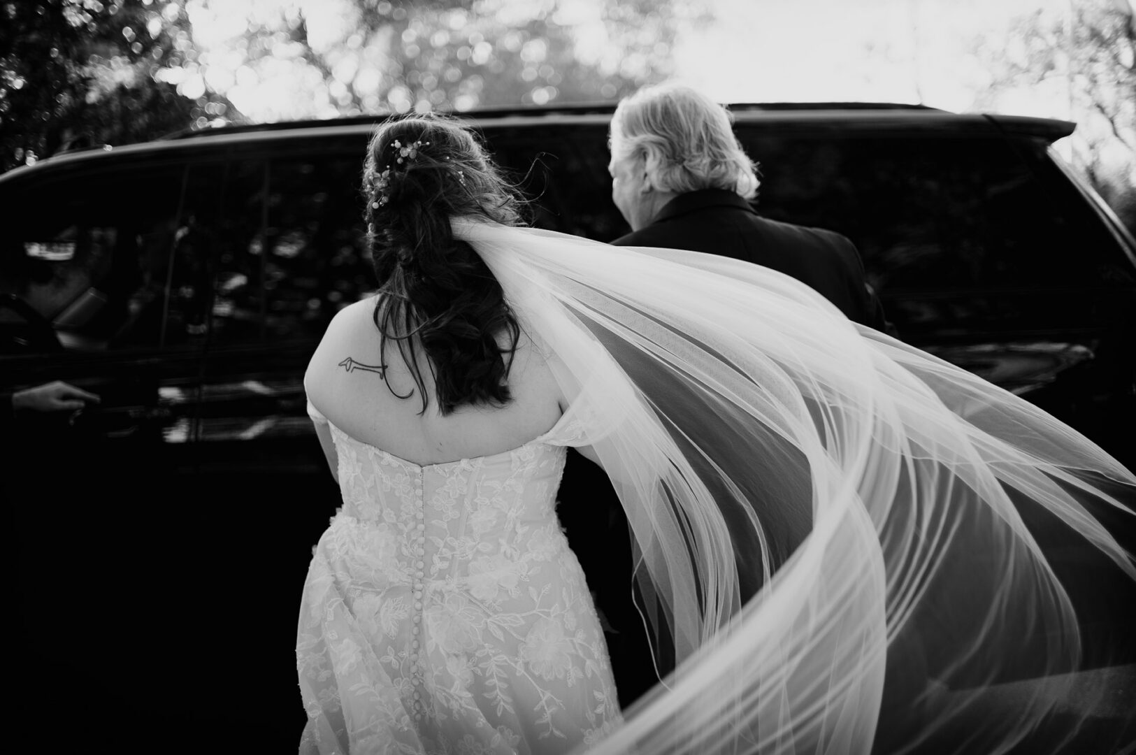 Black-and-white image of the bride entering a car with her veil trailing behind.