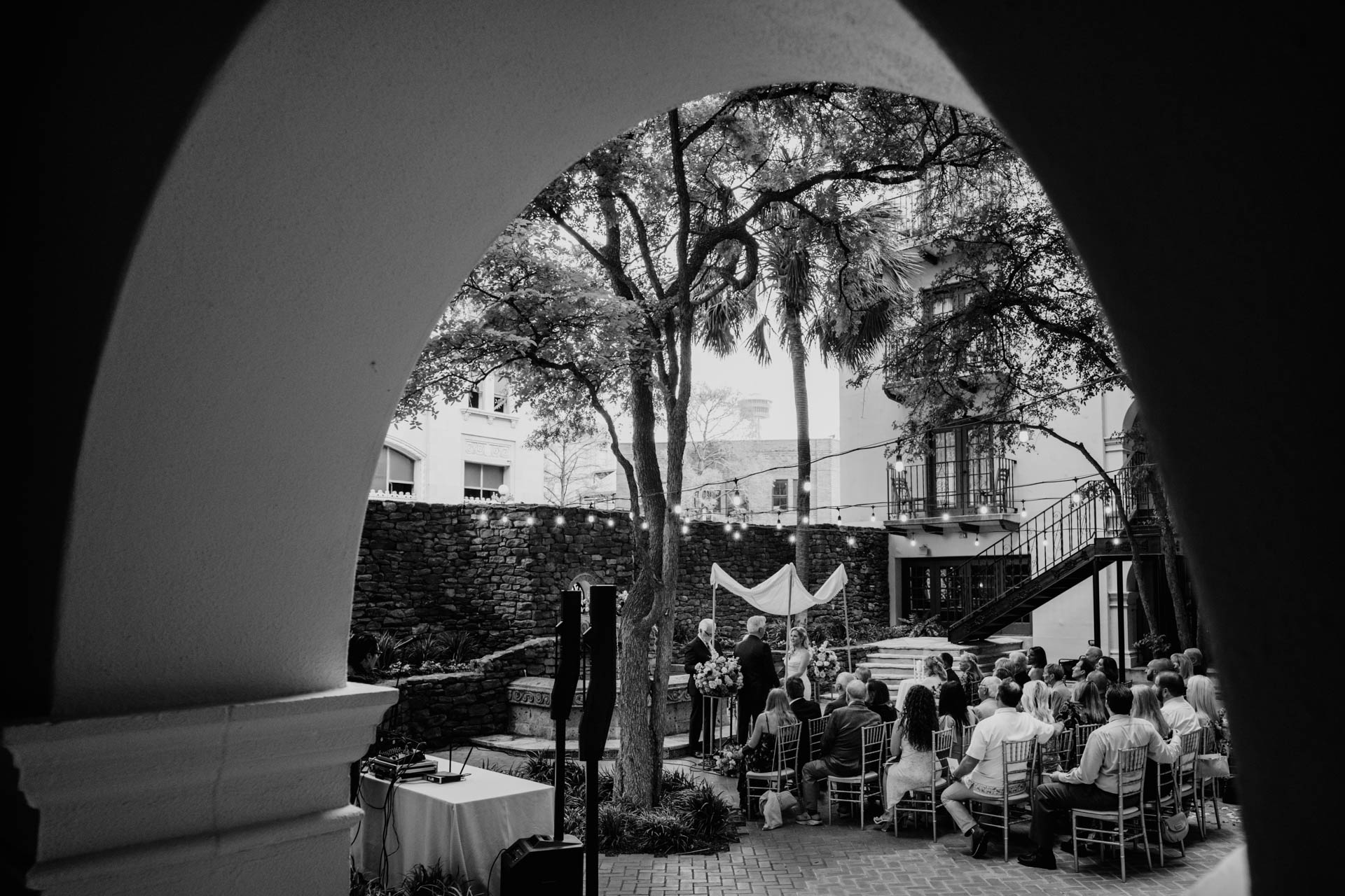 Black and white wide ceremony scene framed through courtyard arch at Omni La Mansión del Rio