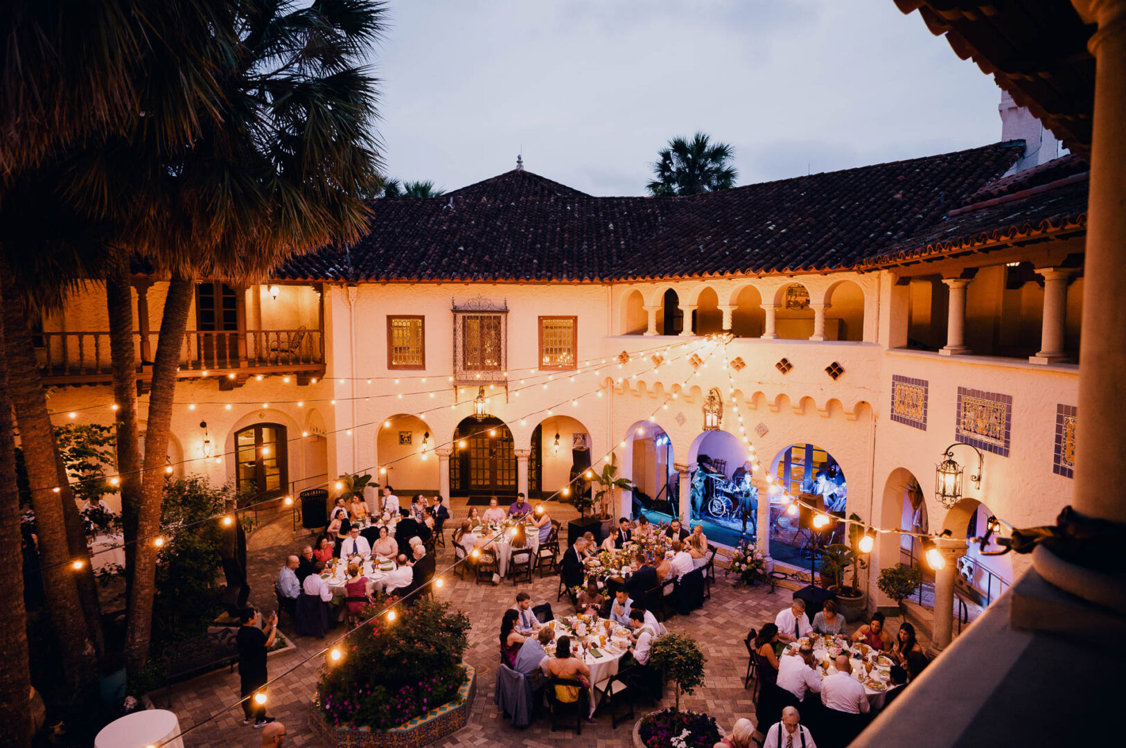 Wide evening view of a historic courtyard wedding reception with guests and string lights.
