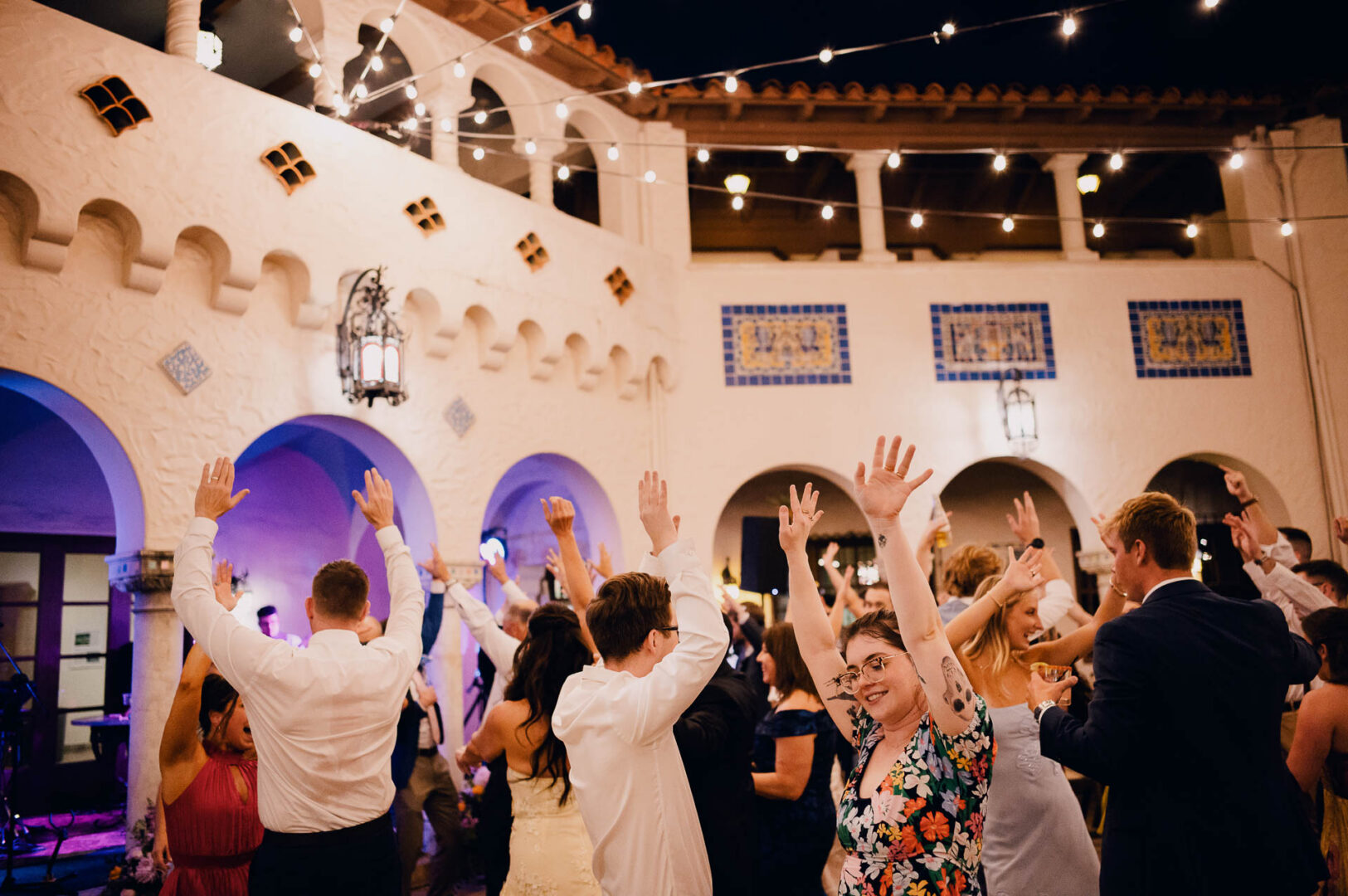 Wedding guests cheering and dancing beneath string lights in a historic courtyard.