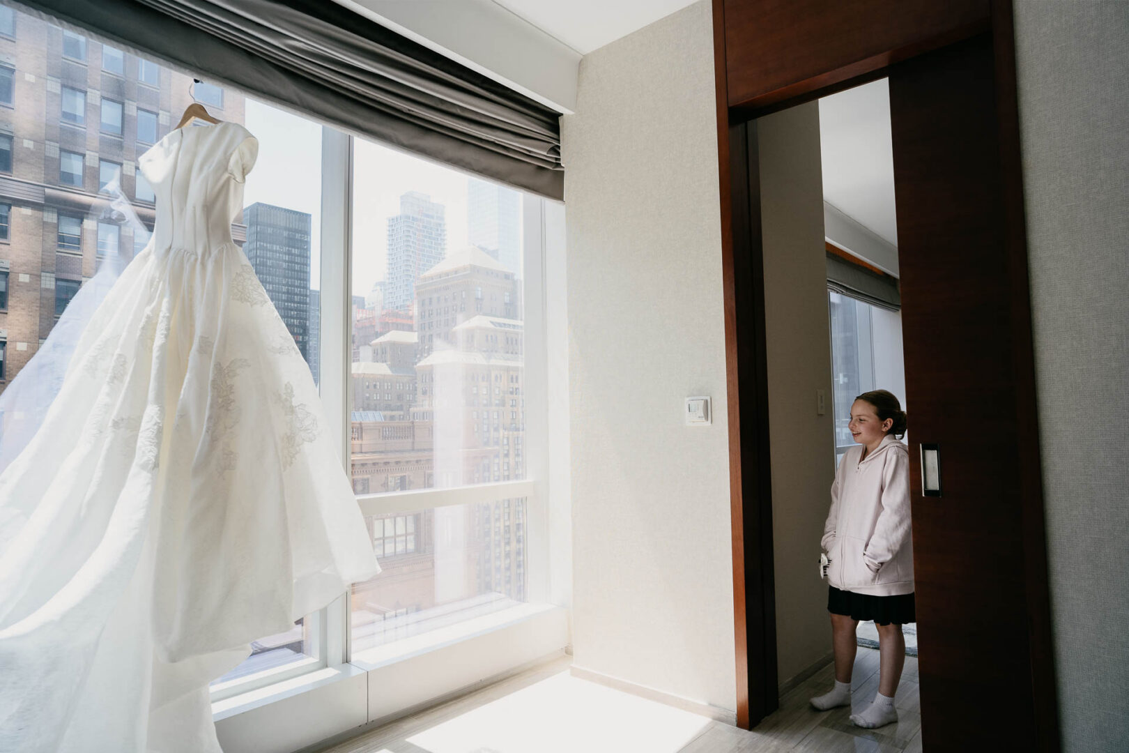 Flower girl standing near a hotel window with the New York skyline in the background.