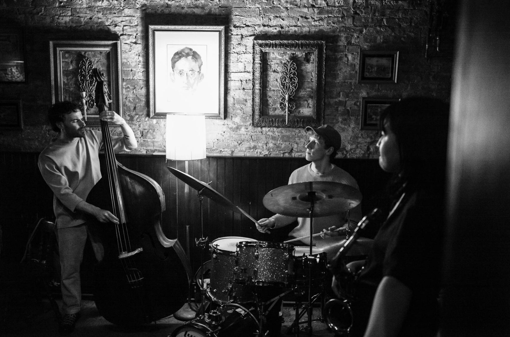 NYC in May: Street Photography in New York City 3 Black-and-white photograph of a jazz trio performing in an intimate New York City bar, with an upright bassist, drummer, and saxophonist framed by a brick wall and lamplight.
