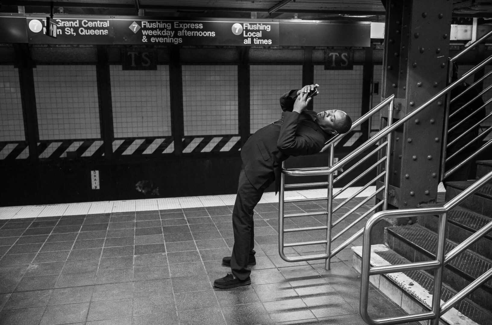 received my first ever Leica Mastershot. Thank you, LFI - Leica Fotografie International - Black-and-white street photograph of a man leaning back against a subway railing while looking at his phone on the 7 train platform at Times Square–42nd Street in New York City.