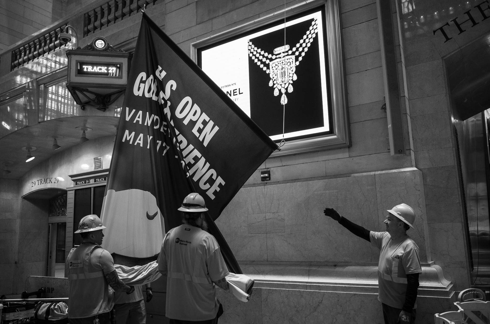 Street + urban photography 132 Black-and-white photograph of workers in hard hats unfolding a large banner near Track 27 inside Grand Central Terminal in New York City.