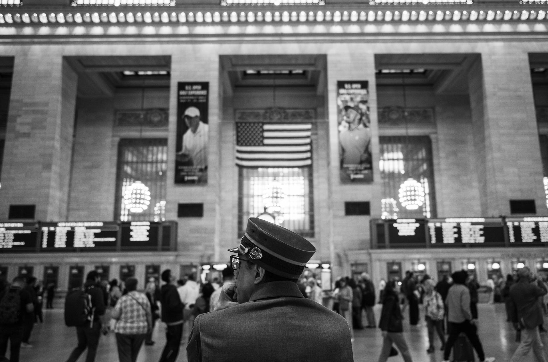 Street + urban photography 133 Black-and-white street photograph of a uniformed worker standing beneath the American flag in the main concourse of Grand Central Terminal as commuters pass through the station.