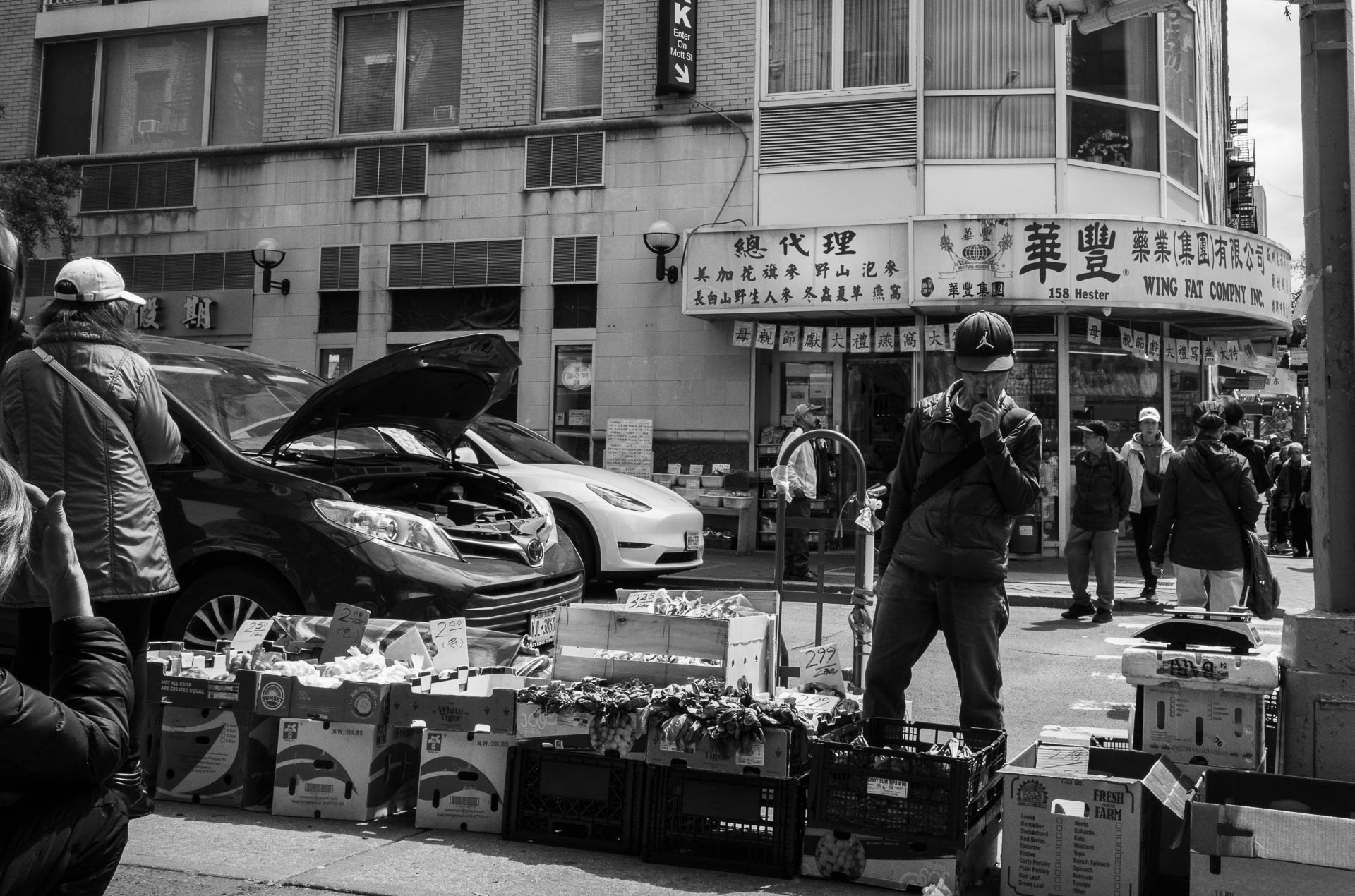 Street + urban photography 136 Black-and-white street photograph of a produce stand on Hester Street in Manhattan’s Chinatown, with pedestrians, parked vehicles, and Chinese storefront signs in the background.