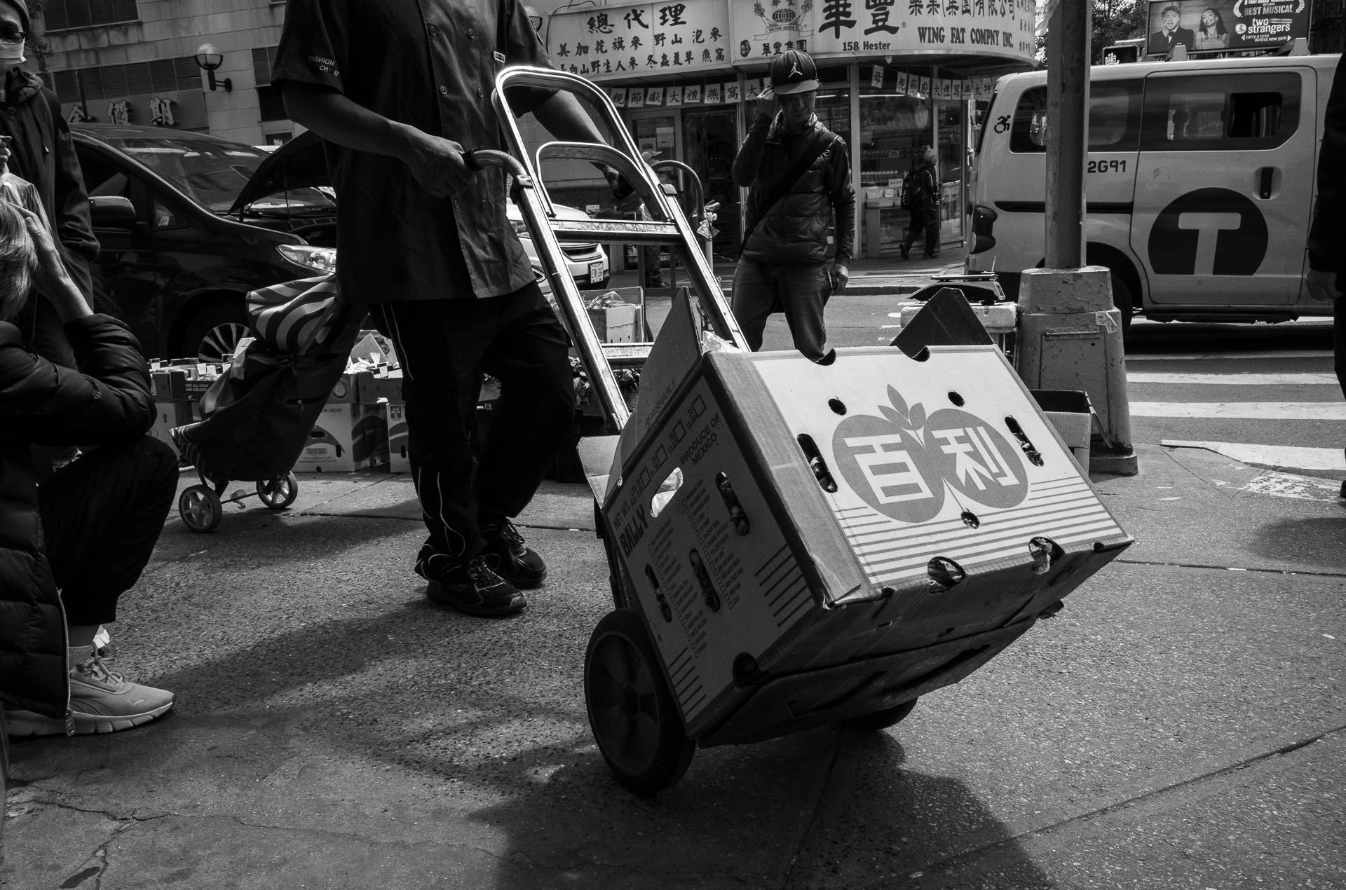 Street + urban photography 137 Black-and-white street photograph of a delivery worker pulling a hand truck loaded with a produce box past a Chinatown sidewalk market in New York City.