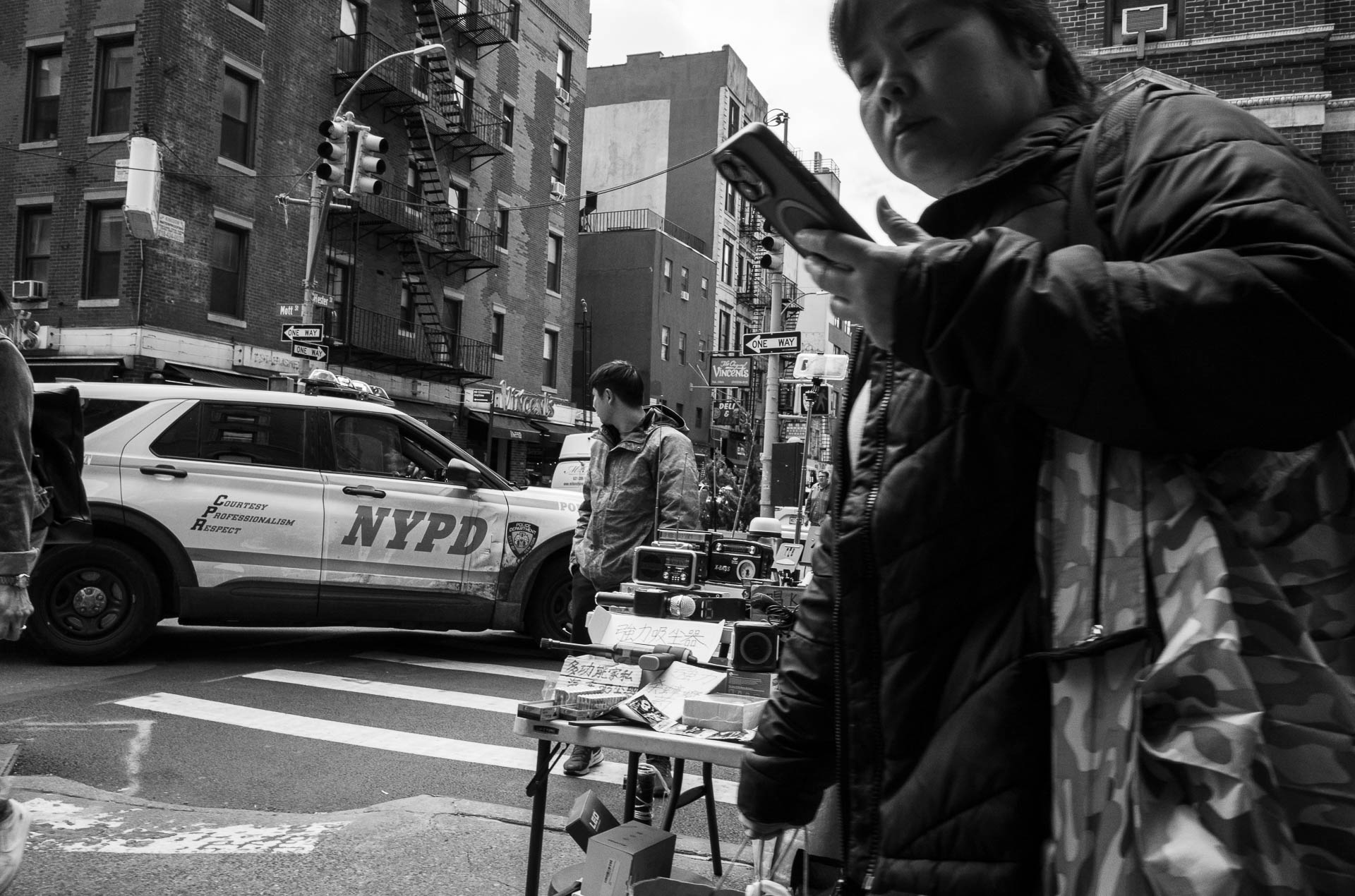 Street + urban photography 138 Black-and-white street photograph of a woman checking her phone near an NYPD vehicle and a sidewalk vendor on Mott Street in Manhattan’s Chinatown.
