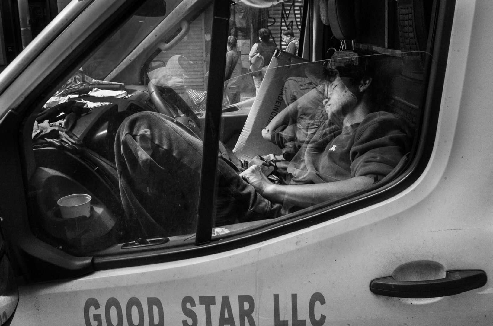 NYC in May: Street Photography in New York City 14 lack-and-white street photograph of a delivery driver resting inside a van, photographed through the window with layered reflections from a Chinatown street in New York City.