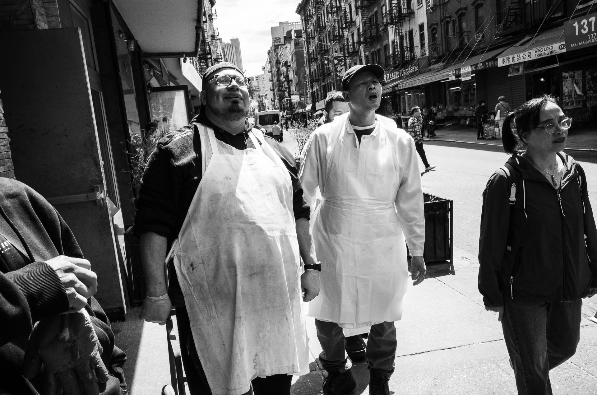 Street + urban photography 139 Black-and-white street photograph of two workers wearing aprons standing outside a Chinatown storefront as pedestrians pass along the sidewalk.