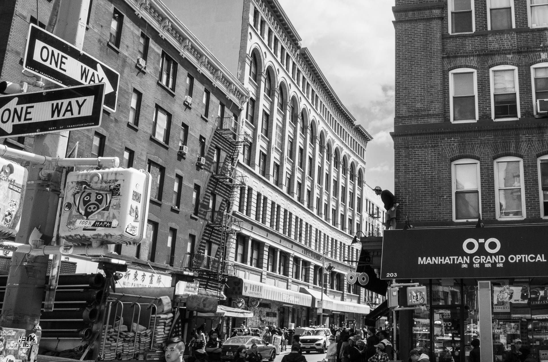 Street + urban photography 141 Black-and-white street photograph of a busy Manhattan Chinatown intersection with one-way signs, fire escapes, storefronts, pedestrians, and a worker standing above an optical shop awning.