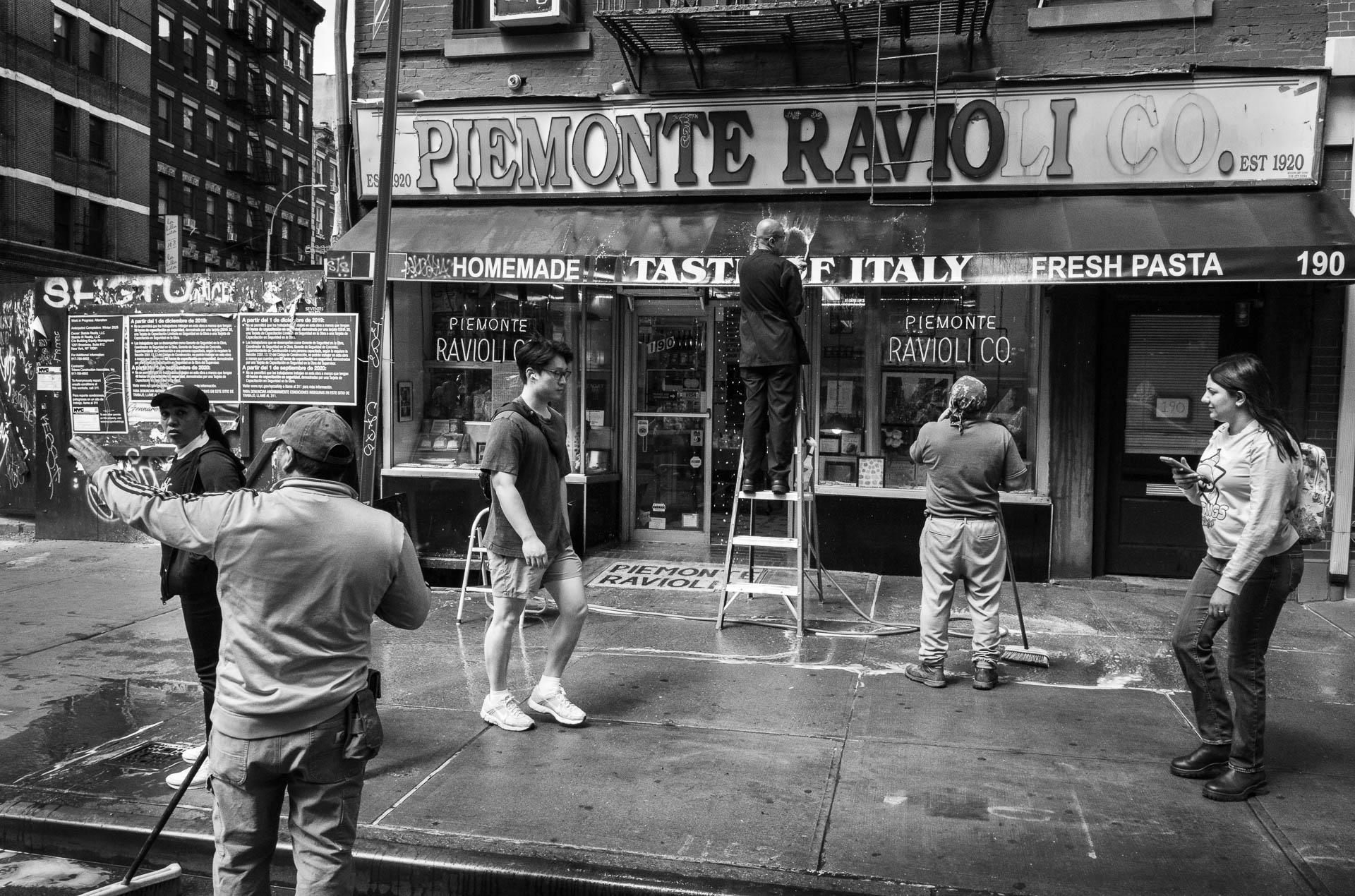 Street + urban photography 142 Black-and-white street photograph of workers washing the storefront and awning of Piemonte Ravioli Co. in Manhattan’s Little Italy as pedestrians pass on the sidewalk.