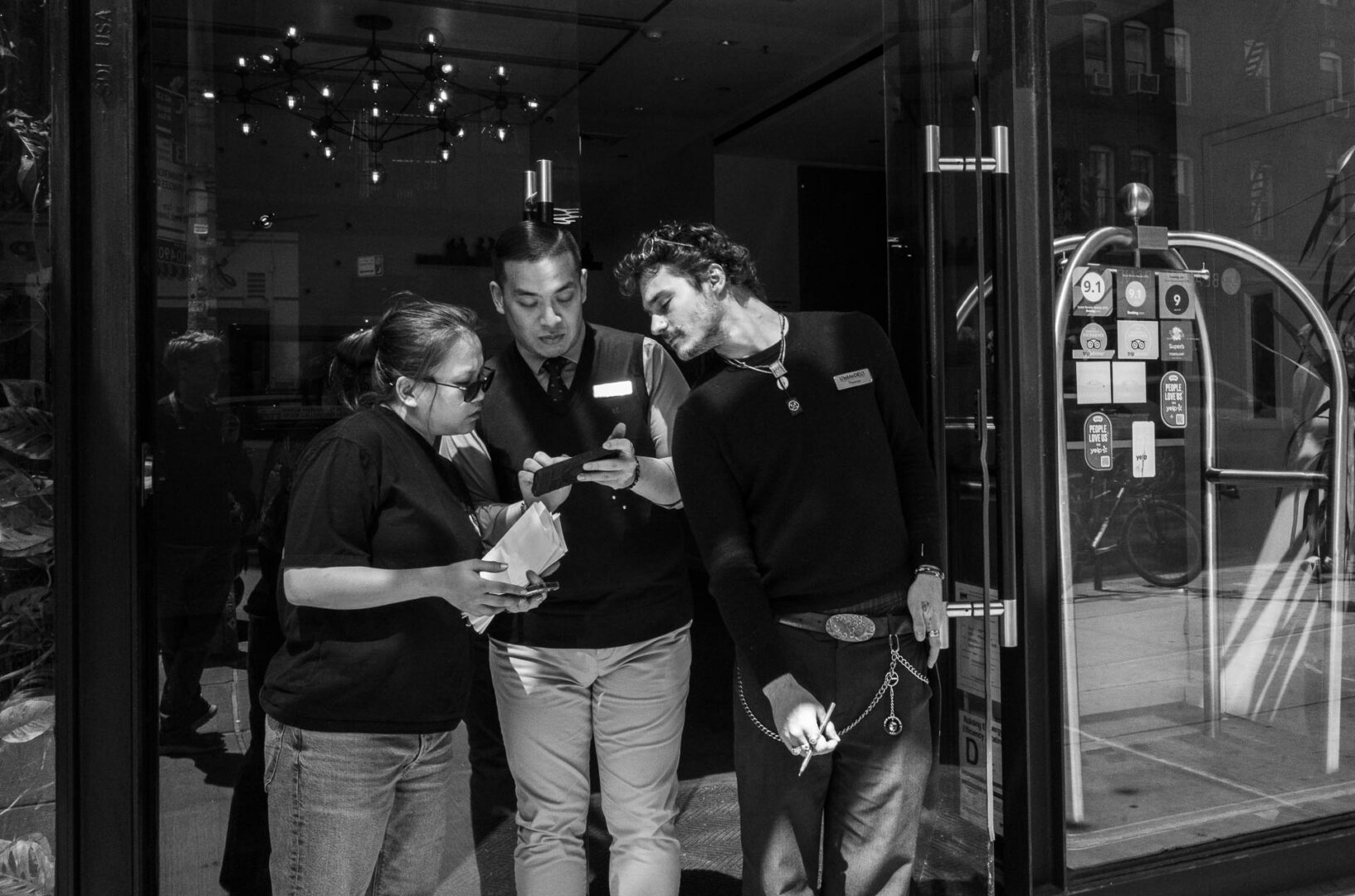 NYC in May: Street Photography in New York City 19 lack-and-white street photograph of three people gathered outside a hotel entrance, looking closely at a phone in the midday light.