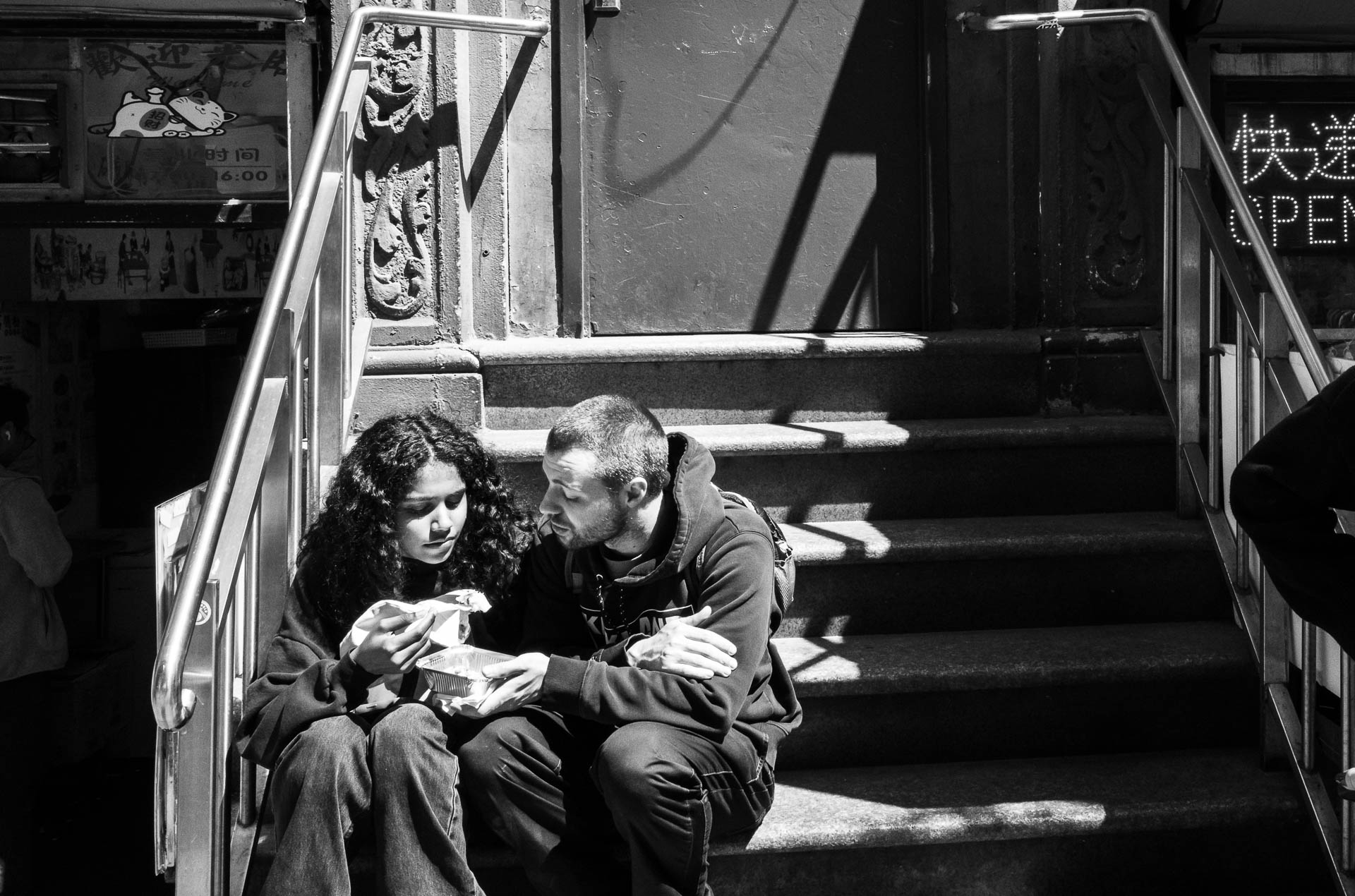 Street + urban photography 7 Black-and-white street photograph of a couple sitting closely together on the steps of a Chinatown storefront while sharing a takeaway meal.